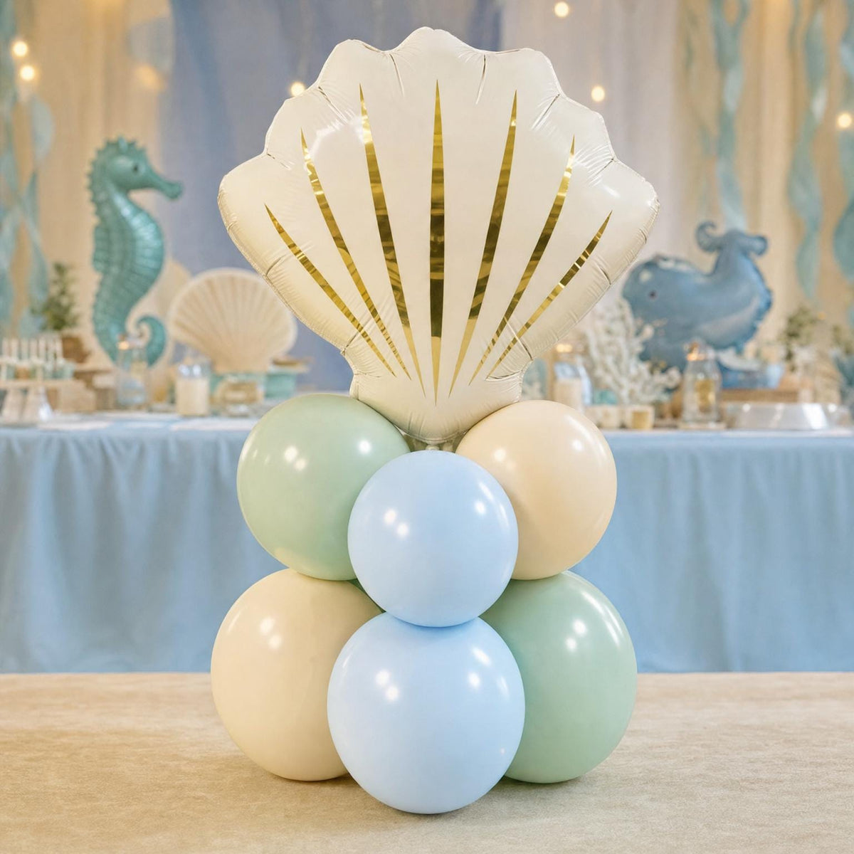 A seashell-shaped balloon cluster with gold foil accents and six pastel balloons in light blue, mint green, and cream, displayed on a table in a decorated party setting.